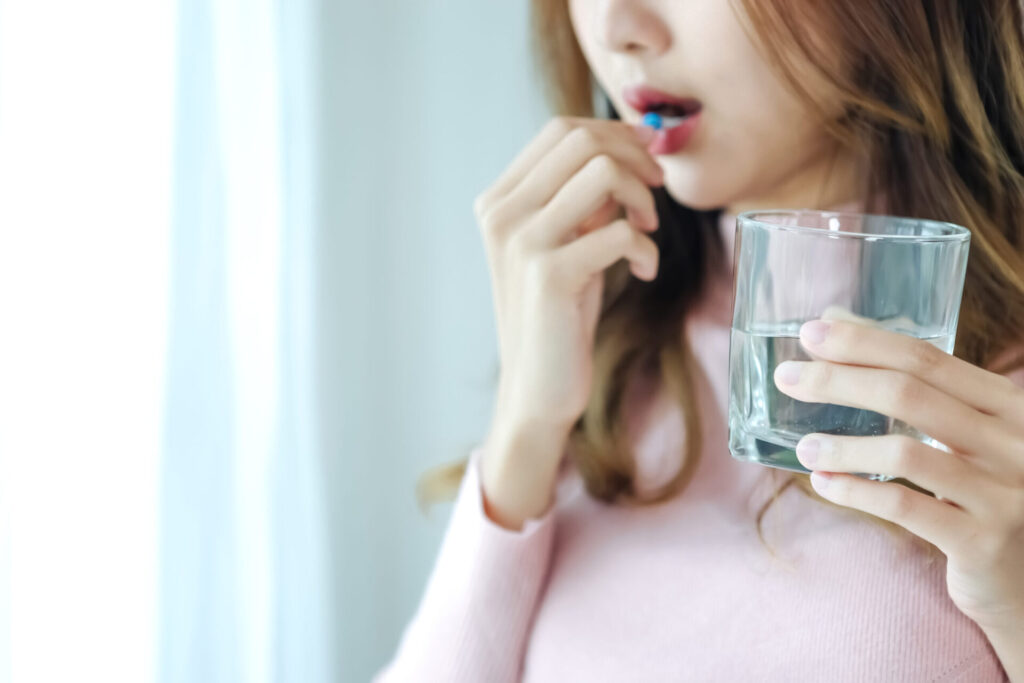 Sick Asian woman eating pills with a glass of water in hand near window in her house.