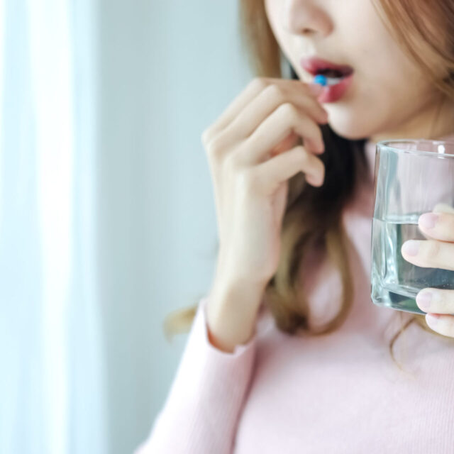 Sick Asian woman eating pills with a glass of water in hand near window in her house.
